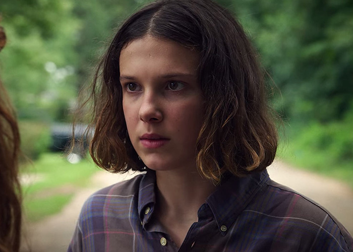 Millie Bobby Brown en camisa de cuadros al aire libre, expresión concentrada, icono clave de Stranger Things.