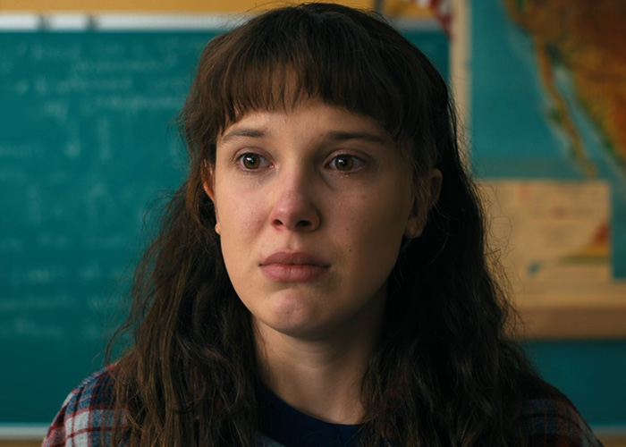 Millie Bobby Brown en primer plano con expresión emocional en escena icónica de Stranger Things en aula escolar.