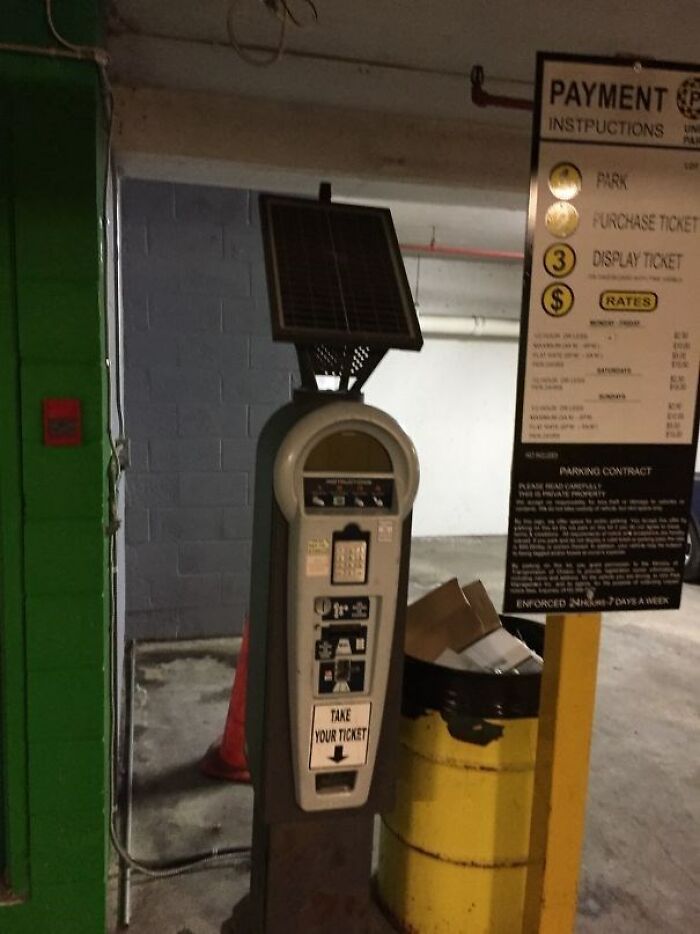 Máquina de tickets solar en estacionamiento con cartel de instrucciones y bote de basura, objeto inútil y divertido sin razón.