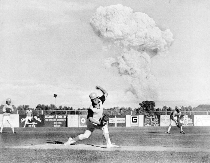 Jugador de béisbol lanzando mientras una enorme nube de explosión ocurre cerca, imagen histórica que no veremos en la vida real.