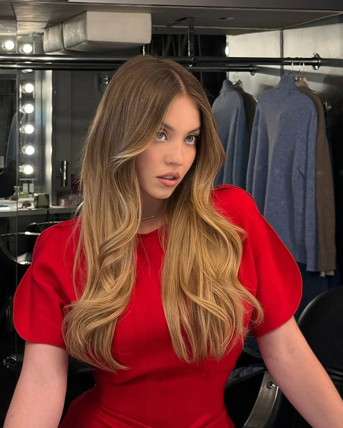 Sydney Sweeney mostrando su belleza natural con cabello peinado y maquillaje en un vestidor con ropa de fondo.