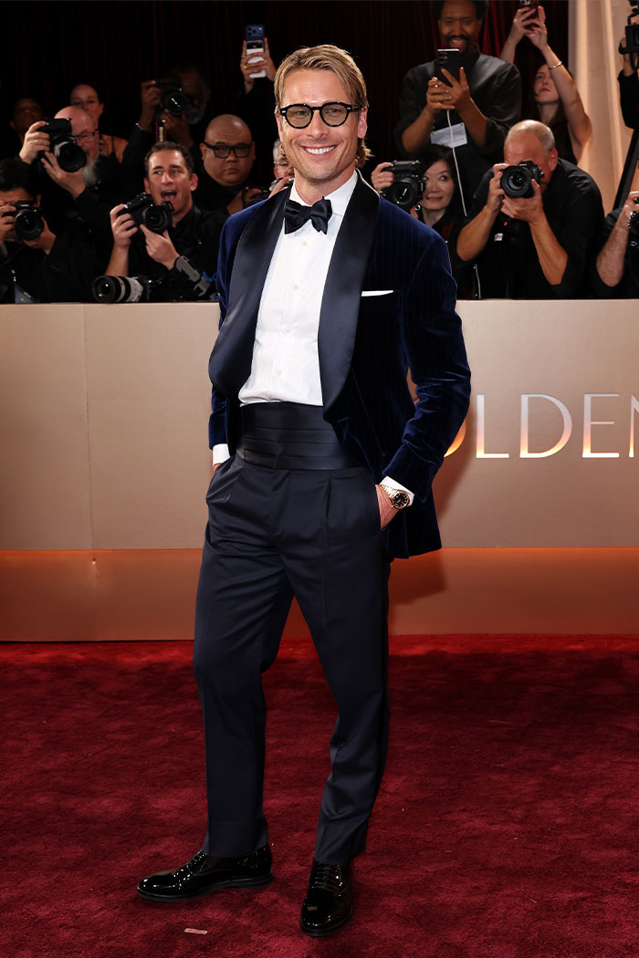 Hombre sonriente en traje elegante azul oscuro con pajarita en la alfombra roja de los Golden Globes, rodeado de fotógrafos.