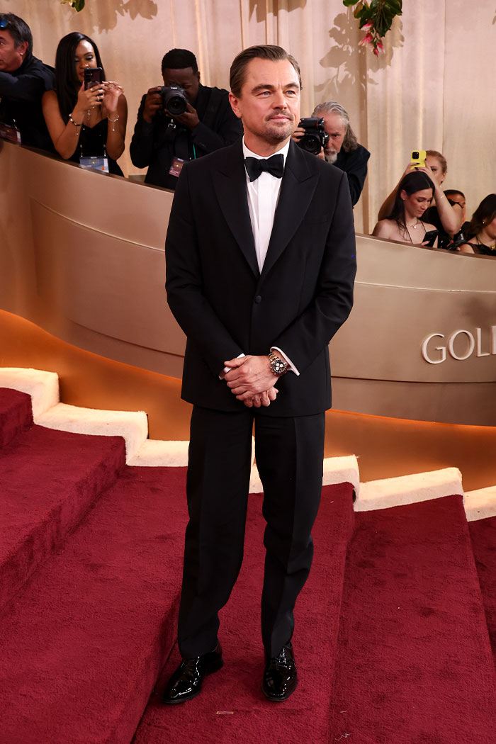 Hombre en esmoquin negro posando en alfombra roja con estilo elegante en evento de Golden Globes con atuendo llamativo.