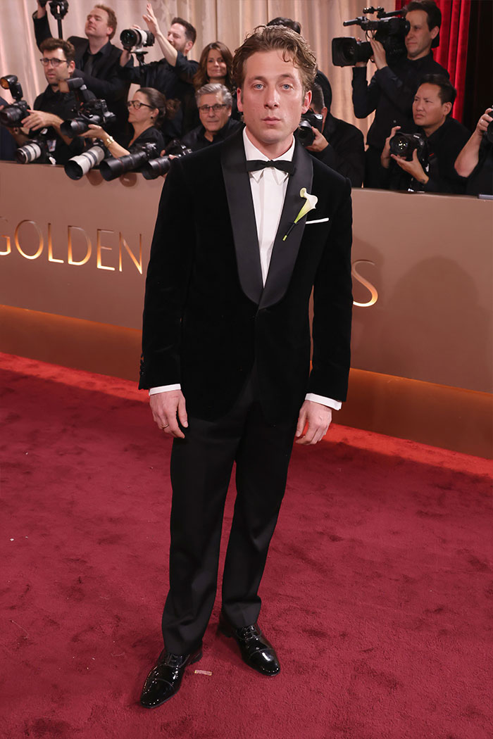 Hombre vestido con esmoquin negro elegante en la alfombra roja de los Golden Globes con fotógrafos al fondo.