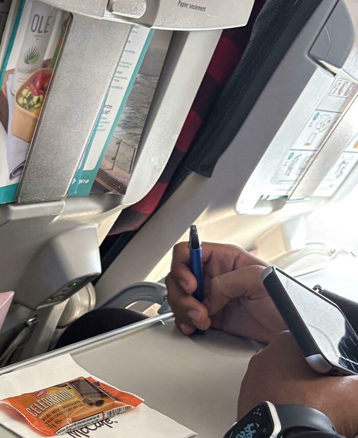 Persona escribiendo con bolígrafo y usando el móvil, en un asiento de avión ignorando a otros pasajeros molestos online.