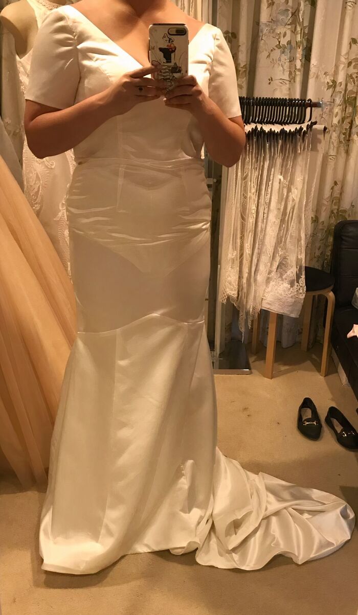 Novia probándose vestido de boda blanco con cola larga en probador junto a otros vestidos y zapatos negros.