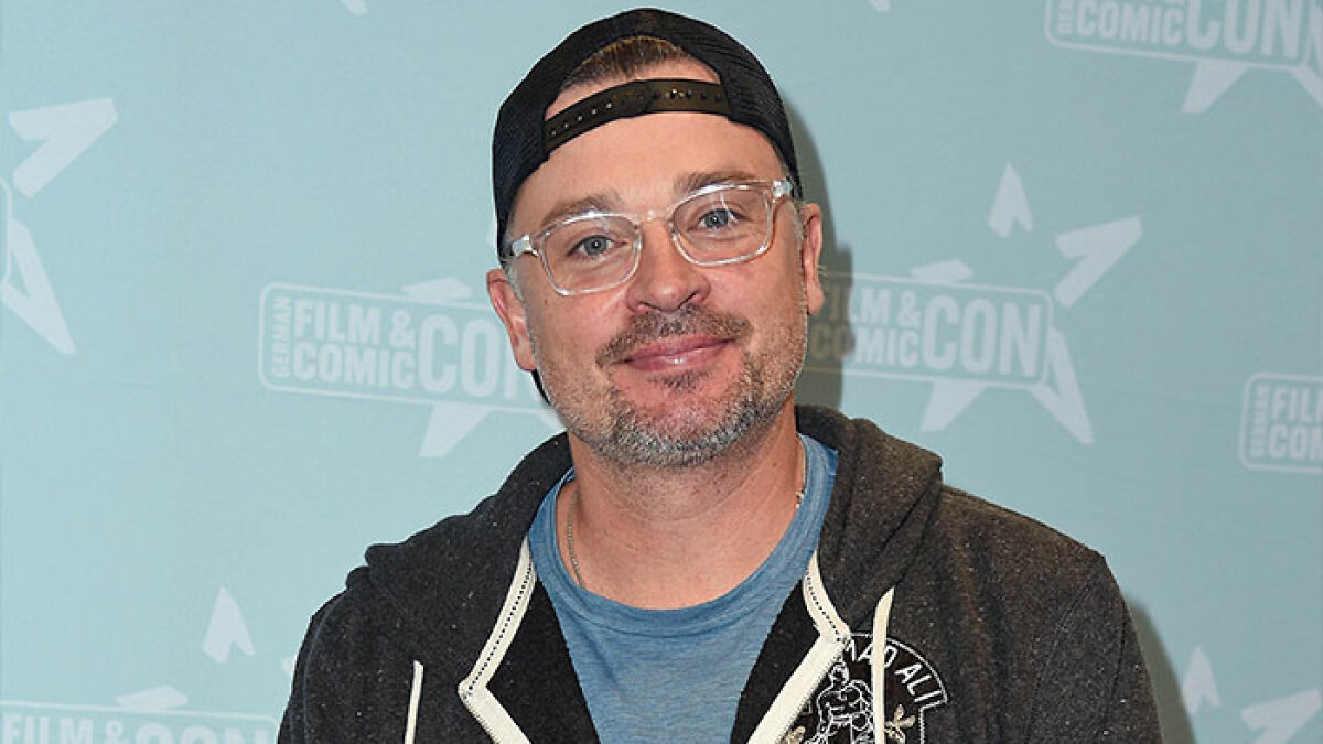 Hombre de 48 au00f1os, galu00e1n adolescente de los 2000, con gorra y gafas transparentes, sonriendo en evento Comic Con.
